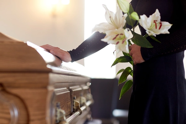 A woman gently holds a white lily in front of a wooden casket, symbolizing remembrance and tranquility. A woman gently holds a white lily in front of a wooden casket, symbolizing remembrance and tranquility.